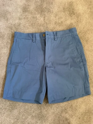 Blå shorts från Polo Ralph Lauren - Snygga blå shorts från Polo Ralph Lauren med stretch och rak passform. Klassisk design med bälteshällor, sidofickor och dragkedja. Perfekta för varma dagar och enkel att matcha med t-shirt eller skjorta. Shortsen är aldrig använda då dem inte passar mig, så endast legat i garderoben.