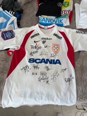 Vit och röd Assyriska FF matchtröja från Allsvenskan - Säljer en äkta använd vit och röd matchtröja från Assyriska FF år 2005 allsvenskan. Tröjan har nummer 16 på ryggen och är signerad av flera spelare. Klassisk fotbollströja i polyester med klubbmärke på bröstet. Perfekt för assyrier som är stolta över sitt lag att hänga upp!