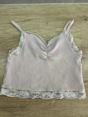 Ljusrosa ribbad linnetopp med spets - Supersöt ljusrosa ribbad linnetopp med smala axelband och vit spetskant runt urringning och nederkant. Perfekt för sommaren och enkel att styla med jeans eller kjol. Ger en romantisk och trendig vibe. Storlek:M