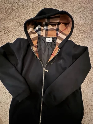 Burberry med dragkedja - Svart hoodie från Burberry med klassiskt rutigt foder i huvan. Dragkedja framtill, långa ärmar och diskret logga på bröstet. Perfekt för en stilren och trendig look. Mjuk bomullskänsla och snygga detaljer på snörena. Fraktas samma dag eller dagen efter!
