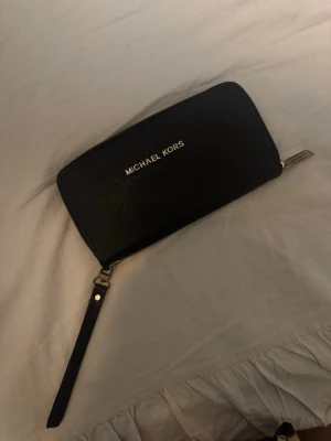 Svart plånbok Michael Kors - Snygg svart plånbok från Michael Kors med guldfärgade detaljer och dragkedja runtom. Plånboken har flera fack för kort och mynt, samt ett handledsband. Insidan är fodrad med MK-logga i beige och guld. Perfekt för att hålla ordning på dina grejer.