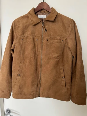Brun mockaimitationsjacka  - Snygg brun jacka i mockaimitation från H&M med dragkedja framtill, två bröstfickor och två sidofickor med knapp. Klassisk krage och raka ärmar ger en stilren look. Perfekt för dig som gillar tidlös och enkel stil.