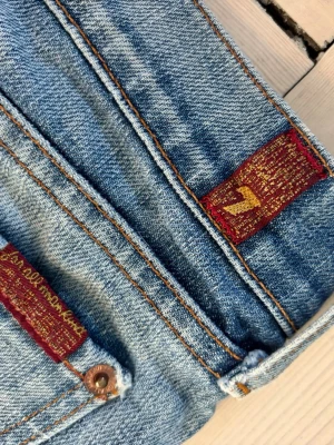 Bootcut jeans från 7 For All Mankind - Snygga blå bootcut jeans från 7 For All Mankind med klassisk femficksdesign och detaljer i rött och guld på etiketten. Jeansen har en lätt tvättad look och subtila slitningar för en avslappnad stil. Storlek 25 men snarare 24. 