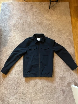 Mörkblå overshirt med fickor - Stilren mörkblå overshirt med dragkedja framtill och två bröstfickor med lock och knapp. Klassisk krage och raka ärmar. Perfekt lager-på-lager-plagg för en clean look.  Nypris 1200