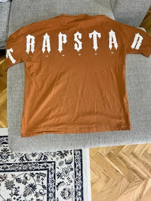Trapstar London brun t-shirt - Snygg brun t-shirt från Trapstar London med vit logga tryckt både fram och bak. T-shirten har en klassisk passform och är tillverkad i mjuk bomull, perfekt för en avslappnad streetwear-look.