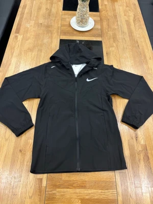 Svart vindjacka från Nike med huva - Svart vindjacka från Nike med hel dragkedja och huva. Jackan har en stilren design med Nike-logga på bröstet och elastiska muddar vid ärmsluten. Perfekt för blåsiga dagar och sportiga outfits.