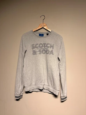 Grå sweatshirt från Scotch & Soda - Grå sweatshirt med tryck 'Scotch & Soda Amsterdams Blauw' på bröstet. Tröjan har rund halsringning, långa ärmar och ribbade muddar med svarta ränder. Perfekt för en avslappnad och stilren look.