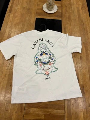 Vit Casablanca Paris t-shirt - Vit t-shirt från Casablanca Paris med färgglatt tryck på ryggen och mindre tryck framtill. Klassisk passform och rund hals. Perfekt för dig som gillar stilrena plagg med unika detaljer.