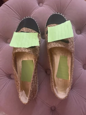 Glittriga guldiga espadrille-sandaler - Säljer ett par espadrille-sandaler i glittrigt guldtyg med svart tåparti och flätad sula i naturmaterial. Skorna har öppen design och är perfekta för dig som vill ha något extra till sommaren. Snygg kontrast mellan guld och svart.