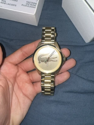 Guldfärgad klocka från Lacoste - Stilren guldfärgad klocka från Lacoste med metallarmband och stor krokodil-logga på urtavlan. Klockan har en rund form och klassisk design som sticker ut med sin eleganta finish. Perfekt accessoar för dig som vill ha något extra.