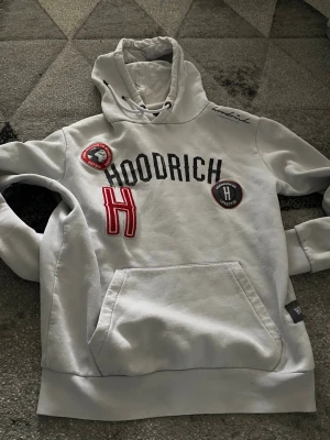 Vit hoodie från Hoodrich med patchar - Snygg vit hoodie från Hoodrich med broderade patchar och tryck på bröstet. Stor röd H, svarta bokstäver och coola detaljer. Mjuk insida och klassisk känguruficka framtill. Perfekt för en streetwear-look.