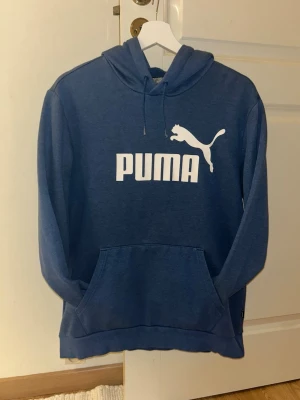 Blå hoodie från Puma med logga - Snygg blå hoodie från Puma med stor vit logga och text på bröstet. Klassisk känguruficka framtill och justerbar huva med dragsko. Perfekt för en avslappnad och sportig stil. Mjuk och bekväm bomullsblandning. Storlek M