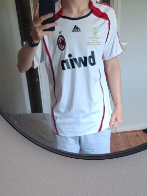 AC Milan Kaká 22 Adidas - Säljer en vit AC Milan bortatröja från Adidas med röda detaljer och tryck. Framsidan har klubbmärke, Adidas-logga och Champions League-märke, samt bwin som sponsor. Baksidan har Kaká och nummer 22 i rött. Perfekt för fotbollsfans!