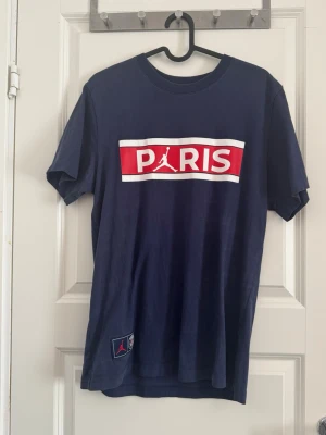 Mörkblå Paris Jordan t-shirt - Snygg mörkblå t-shirt med stort Paris-tryck och Jordan-logga i vitt och rött på bröstet.                           Storlek:S