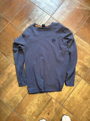 Hugo boss tröja - En enkel och stilren mörkblå långärmad tröja med rund hals och diskret logga på bröstet. Tröjan är tillverkad i mjukt bomullstyg och passar perfekt till jeans eller chinos för en avslappnad look.