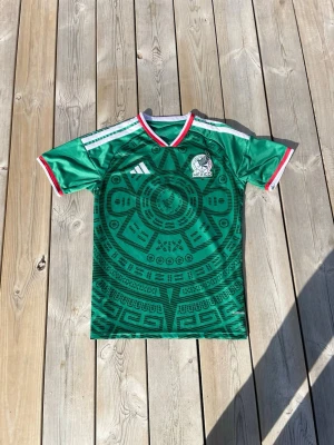 Mexikansk grön fotbollströja L passar M - Snygg grön Mexiko fotbollströja från Adidas med aztekiskt mönster över hela tröjan. Tröjan har vita och röda detaljer på ärmar och krage samt Mexikos landslagsemblem på bröstet. Perfekt för match eller träning.