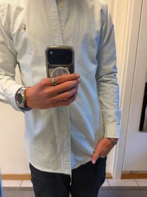 Randig skjorta från Polo Ralph Lauren - Säljer en klassisk ljusgrön och vit randig skjorta från Polo Ralph Lauren i modellen Custom Fit. Skjortan har långa ärmar, knappar framtill och den ikoniska broderade loggan på bröstet. Tillverkad i mjuk bomull som känns skön mot huden. Storlek M