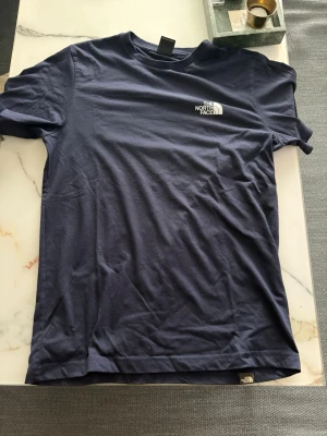 Blå The North Face t-shirt - Säljer en mörkblå t-shirt från The North Face med liten logga på bröstet och stort tryck med texten 'Never Stop Exploring' och bergsmotiv på ryggen. Klassisk passform och rund hals. Perfekt för dig som gillar outdoor-vibbar