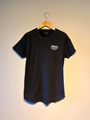 Svart t-shirt från Hollister - Snygg svart t-shirt från Hollister med vit logga och text på bröstet. Klassisk rund halsringning och korta ärmar. Perfekt basplagg i mjuk bomull som funkar till allt och ger en clean look.