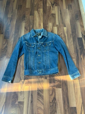 Blå jeansjacka från Lee, storlek S - Klassisk blå jeansjacka från Lee i storlek S. Jackan har två bröstfickor med lock och knapp, samt snygga kontrastsömmar. Tidlös modell med knäppning framtill och krage. Perfekt för dig som gillar en avslappnad och cool stil.