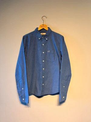Blå skjorta från Hollister - Snygg blå skjorta från Hollister med diskreta tunna ränder och klassisk knappslå. Skjortan har en bröstficka med broderad logga och vita knappar. Perfekt till jeans eller chinos för en clean look.