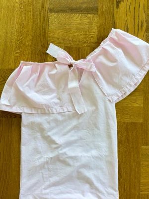 Ljusrosa offshoulder klänning med knyt - Söt ljusrosa klänning i bomull med offshoulder-modell och volang upptill. Klänningen har ett knytband vid axeln som ger en extra detalj. Perfekt för varma dagar och somriga tillfällen. Stor volang med justerbart band skulle säga att den passar en xs/s