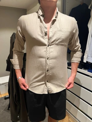Beige flanell skjorta med bröstficka - Stilren beige skjorta med lång ärm och klassisk krage. Skjortan har en bröstficka på vänster sida och knäpps med knappar framtill. Tillverkad i ett mjukt tyg som ger en avslappnad look. Perfekt för dig som gillar enkel och clean stil. Flanell skjorta.