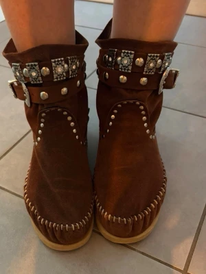 Bruna mocka cowboy boots med nitar - Unika cowboy boots i brun mocka med dekorativa silverfärgade nitar och spännen. Bootsens skaft pryds av mönstrade remmar och detaljerade spännen i westernstil. Sömmen runt sulan är grov och synlig, vilket ger en cool handgjord känsla. Perfekta för dig som vill sticka ut.