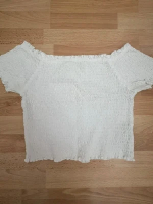 Vit smockad topp med kort ärm - Säljer en söt vit topp med smockat tyg och korta ärmar. Toppen har en lätt croppad passform och elastisk struktur som ger en bekväm känsla. Perfekt till jeans eller kjol för en fräsch look.