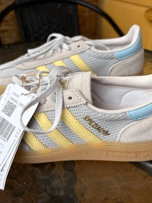 Adidas Handball Spezial sneakers - Adidas Handball Spezial sneakers i ljusgrå mocka och mesh med gula detaljer och ljusblå häl. Klassiska tre ränder på sidan och gummisula. Guldfärgad Adidas-logga och texten 'Spezial' på sidan. Skön retrostil och snygg färgkombo.