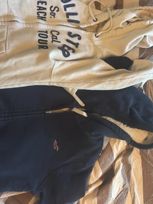 Två hoodies med dragkedja och huva - Säljer två hoodies, en ljus med tryck 'Cali Step So Cal Beach Tour' och en mörkblå med liten broderad logga på bröstet. Båda har dragkedja, fickor och huva med snören. Den mörkblå har mysigt foder i huvan. Perfekta för chill dagar.