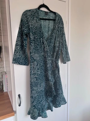 Mörkgrön omlottklänning med trekvartsärm - En tunn klänning med trekvartsärm och omlottdetalj med knytband. Mörkgrön med vitt mönster från Vero Moda. Använd en gång.  Originalpris: 349kr