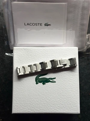 Silverfärgat armband från Lacoste - Snyggt armband från Lacoste i silverfärgad metall med länkdesign och detaljerade mönstrade länkar. Perfekt accessoar för dig som gillar stilrena och sportiga smycken. Levereras i originalask med Lacoste-logga.