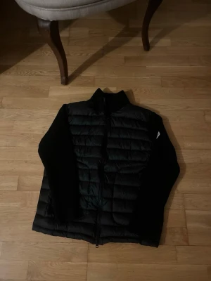 Svart pufferjacka från moncler med stickade ärmar - Säljer en svart pufferjacka med quiltad front och rygg samt stickade ärmar och sidor. Jackan har hög krage och dragkedja framtill. Insidan har en innerficka och ett färgglatt tryck. Perfekt för kalla vinterdagar.