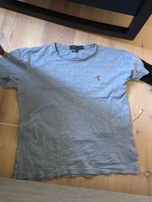 Grå t-shirt från Polo Ralph Lauren - Snygg grå t-shirt från Polo Ralph Lauren med klassisk rund halsringning och den ikoniska broderade loggan på bröstet. T-shirten har korta ärmar och är tillverkad i mjukt bomullsmaterial som känns skönt mot huden.