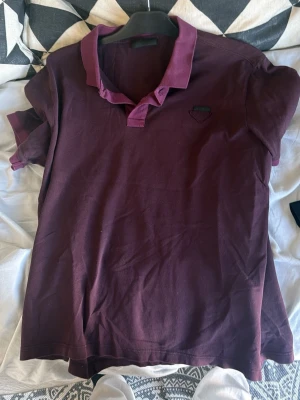 Vinröd pikétröja med krage från Prada  - Säljer en vinröd pikétröja med klassisk krage och korta ärmar. Tröjan har en liten diskret detalj på bröstet och är tillverkad i mjukt bomullstyg. Perfekt för en avslappnad men ändå stilren look.