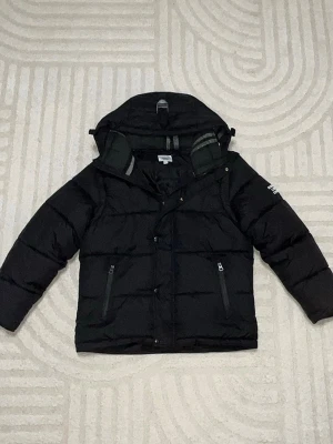 Svart pufferjacka från Burberry - Snygg svart pufferjacka från Burberry med huva och två dragkedjefickor framtill. Jackan har Burberry London England-tryck på ena ärmen och quiltad design. Perfekt för kalla vinterdagar och riktigt trendig look.