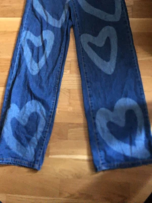 Blå jeans med hjärtmönster - Unika blå jeans med stora, ljusa hjärtan tryckta längs benen. Jeansen har rak passform och är tillverkade i klassiskt denimtyg. Perfekta för dig som vill sticka ut med ett lekfullt och trendigt plagg.