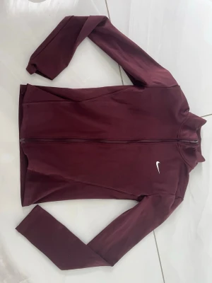 Vinröd zip-jacka från Nike - Snygg vinröd träningsjacka från Nike med hel dragkedja och hög krage. Tillverkad i ett stretchigt funktionsmaterial som passar perfekt till träning eller uppvärmning. Diskret vit Nike-logga på bröstet för en sportig look.