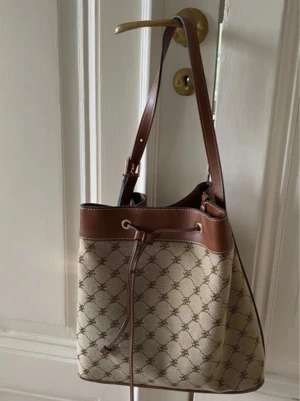 Beige och brun bucket bag med mönster - Snygg bucket bag i beige canvas med brunt läder och justerbar axelrem. Väskan har ett klassiskt monogrammönster och dragskostängning upptill. Perfekt för dig som gillar stilrena detaljer och tidlös design.