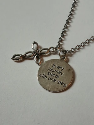 Silverfärgat halsband med berlock - Snyggt silverfärgat halsband med två berlocker: en rund bricka med texten 'Every journey starts with one step' och en dekorativ korsliknande berlock med öppna detaljer. Materialet rostfritt stål.