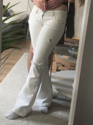 Vita low rise flare jeans - Säljer ett par vita low rise jeans med utsvängda ben och slitningar vid knäna från ginatricot. Jeansen har en klassisk knappgylf. Perfekta för dig som gillar en trendig och avslappnad stil. Användt några gånger, men inget som märkbart syns. Säljes då jag har mina ögon på ett annat par. Första bilden är byxorna på mig. 