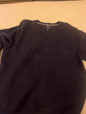 Mörkblå sweatshirt från GANT - Klassisk mörkblå sweatshirt från GANT med rund halsringning och ribbade muddar. Tröjan är enkel och stilren, perfekt för en avslappnad look. Tillverkad i mjukt bomullsmaterial för extra komfort.