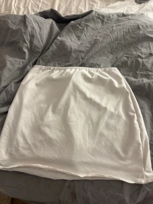 Vit kortkjol med snörning - Säljer en vit kortkjol med snörning där bak. Kjolen har en enkel och clean design med elastisk midja och är gjord i ett mjukt material som känns bekvämt. Perfekt att styla med en crop top eller oversized hoodie.