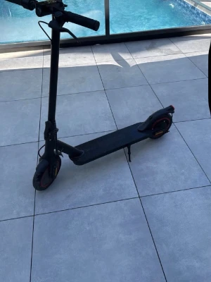 Svart elscooter med röda detaljer - Säljer en svart elscooter med röda detaljer på hjulen. Scootern har en smal, avlång plattform och styre med handtag. Utrustad med stödben och bakre lampa. Perfekt för att ta sig fram snabbt i stan.