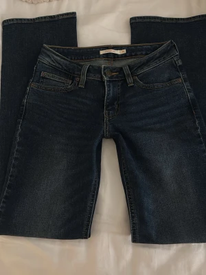 Mörkblå lågmidjade bootcut jeans från Levis - Säljer ett par mörkblå jeans med bootcut-modell och superlow midja. Jeansen har klassiska fem fickor, bälteshällor och snygga slitningar framtill. Perfekta för dig som gillar en avslappnad men trendig look.