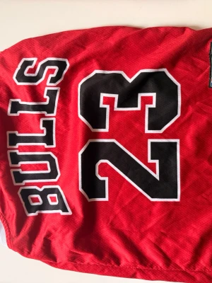 Chicago Bulls basketset röd Jordan 23 - Säljer ett rött basketset med Chicago Bulls tema, tröja och shorts. Tröjan har nummer 23 och namnet Jordan i svart och vitt, samt Bulls-logga och NBA-märke. Shortsen är röda med vita och svarta detaljer och Bulls-logga. Perfekt för basketträning eller streetwear.