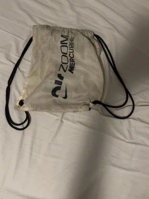 Fotbolls påse - Vit gympåse från Nike med svart Air Zoom Mercurial-tryck och svarta snören. Enkel och smidig design, perfekt för att bära träningskläder eller skor. Tillverkad i lätt tygmaterial och har dragskostängning.