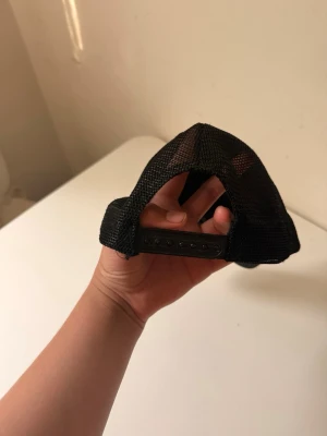 Svart dior keps - Säljer en svart truckerkeps med böjd skärm och justerbar snapback. Kepsen har meshmaterial på sidorna och bak för extra ventilation, perfekt för en avslappnad och sportig stil.