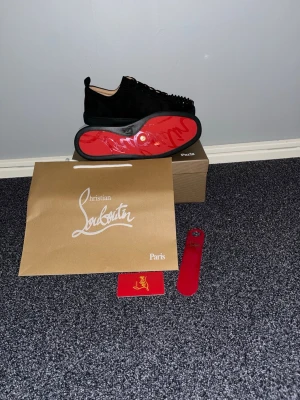 Svarta sneakers med nitar från Louboutin - Säljer ett par svarta sneakers från Christian Louboutin med ikonisk röd sula och svarta nitar över hela ovandelen. Skorna har en låg profil, rund tå och är tillverkade i mocka med läderdetaljer. Grymt statement-plagg för dig som gillar lyx och edgy stil.